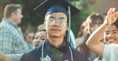Asian graduate in cap and gown, smiling, surrounded by people, outdoor setting.