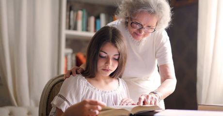 Grandmother helping granddaughter read a book at a desk; sunny interior.