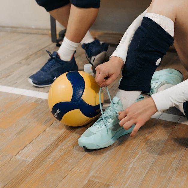 Person tying shoe next to a volleyball on a wooden floor.