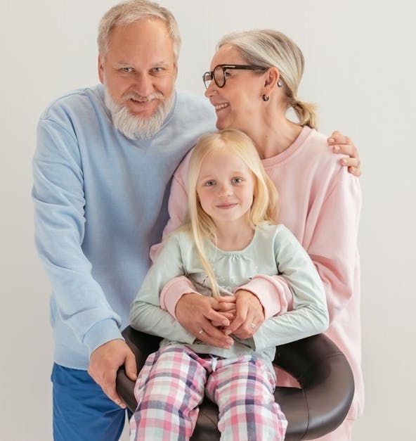 Grandparents embrace a girl in a chair. They smile, wear casual clothes, against a white backdrop.