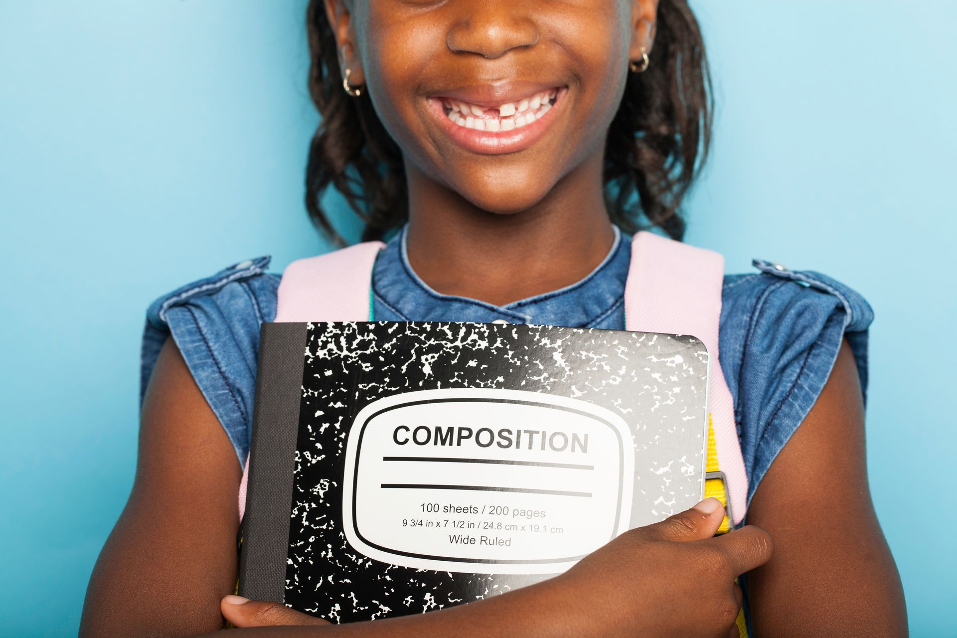 Smiling girl with a composition notebook, backpack, and a missing tooth, against a blue background.
