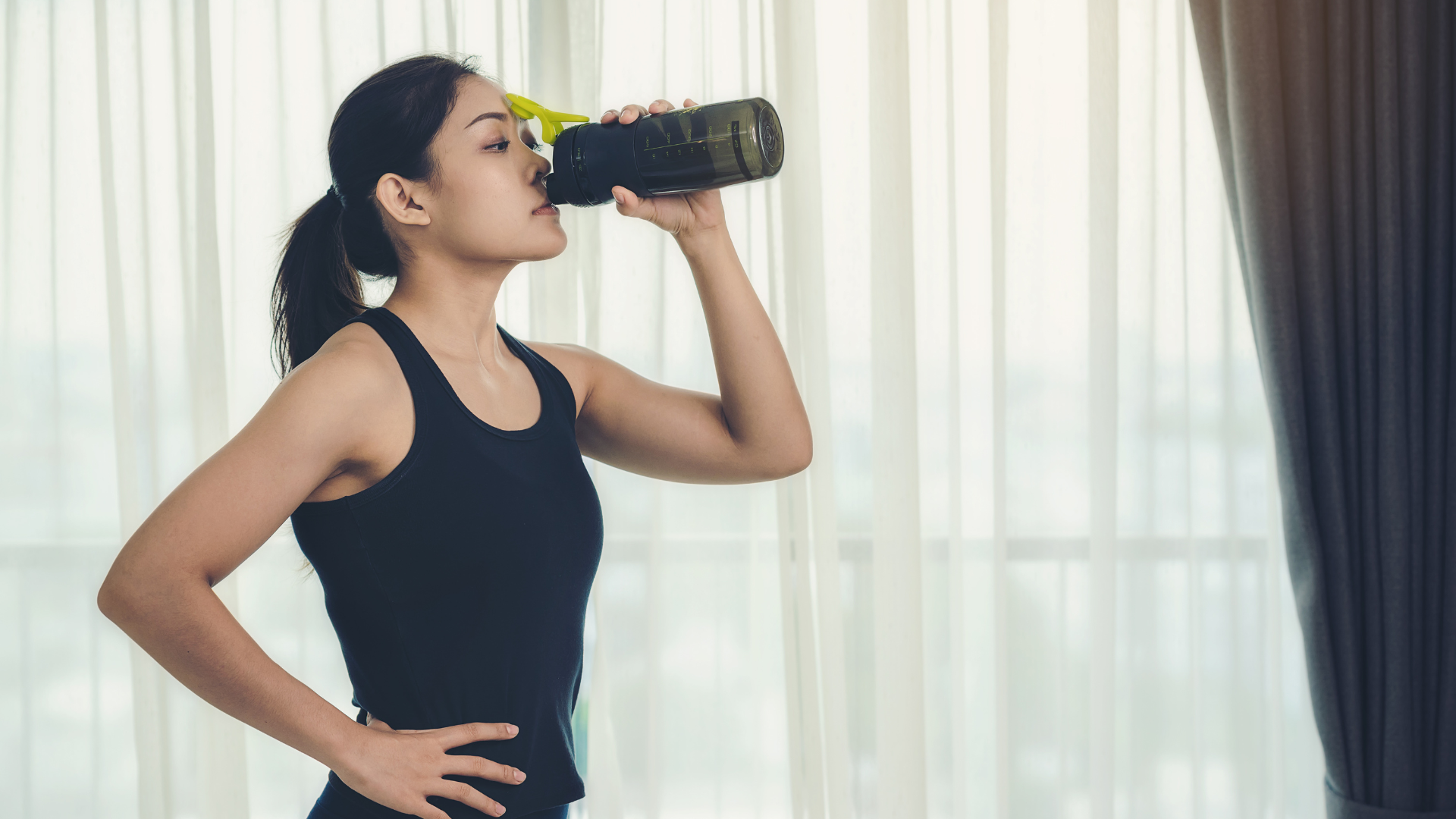 A woman is drinking water from a bottle in front of a window.