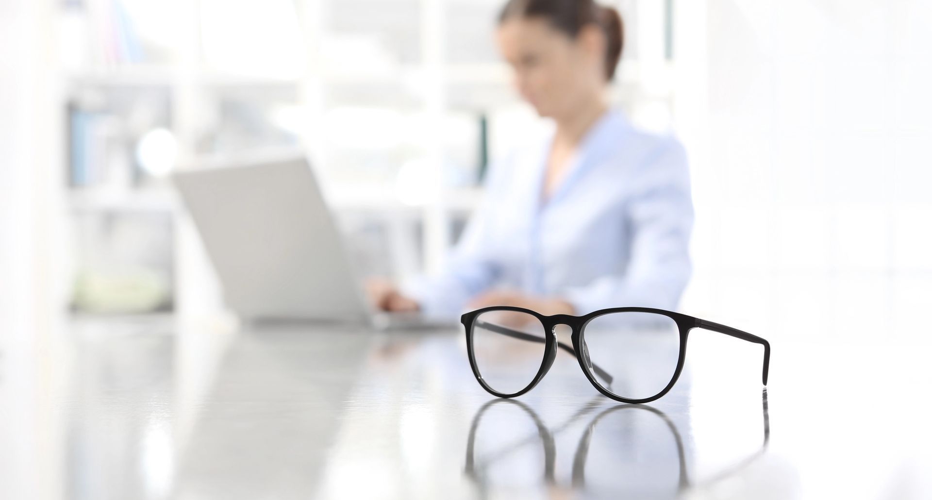 Una mujer está sentada en un escritorio con una computadora portátil y gafas sobre la mesa.