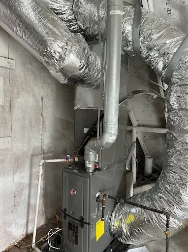 A furnace with ductwork in a utility room, with gray and silver components, against a mottled wall.