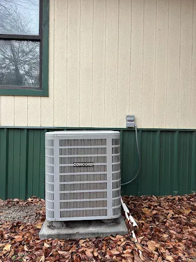 An air conditioning unit outside a building with green and tan siding, beneath a window.