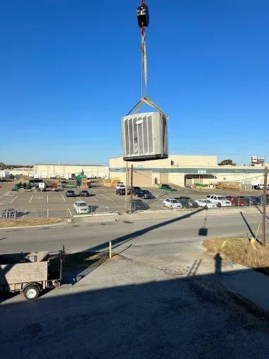 A crane lifts a large, silver industrial unit over an outdoor parking lot on a sunny day.