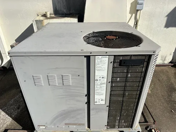 Gray air conditioning unit on a rooftop, with a fan on top and vent on the side.
