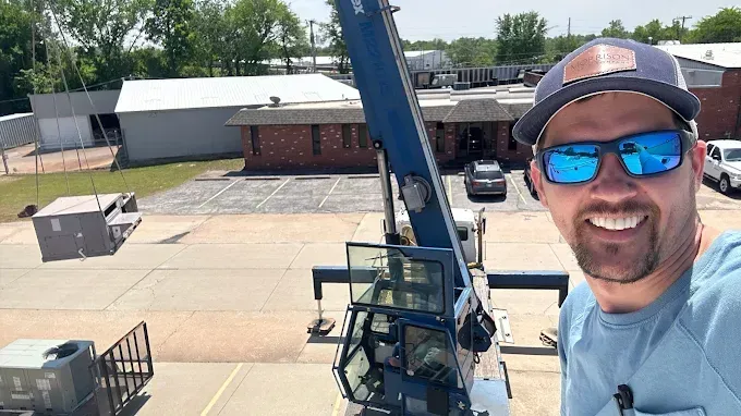 Man in cap and sunglasses smiles at camera from crane cabin. He's lifting equipment onto a building roof.