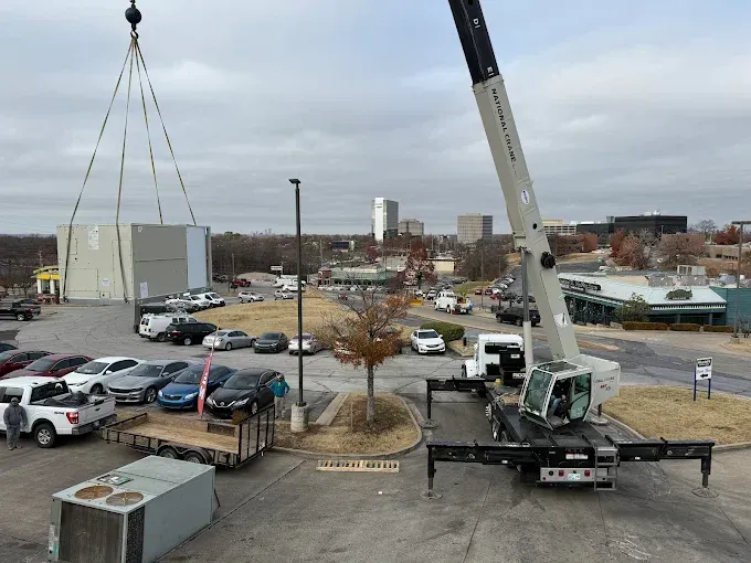 A crane lifting a large rectangular structure, cars parked nearby. City in the background, cloudy sky.