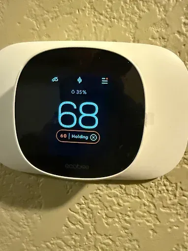 A white thermostat on a tan wall displays a temperature of 68 degrees Fahrenheit. The display also shows the hold setting.