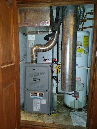 Furnace and hot water heater in a utility closet. Gray metal units with silver pipes against a light blue wall.