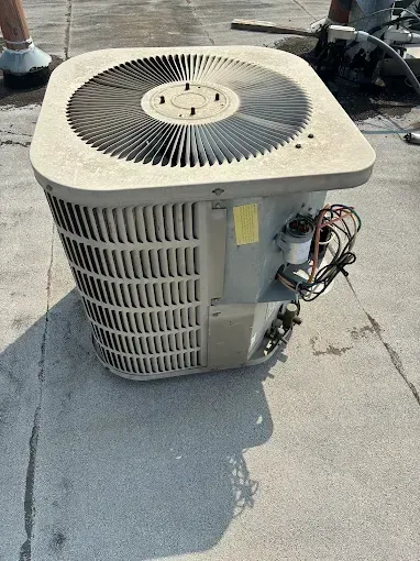 Gray air conditioning unit on a rooftop, with a fan on top and wires visible.