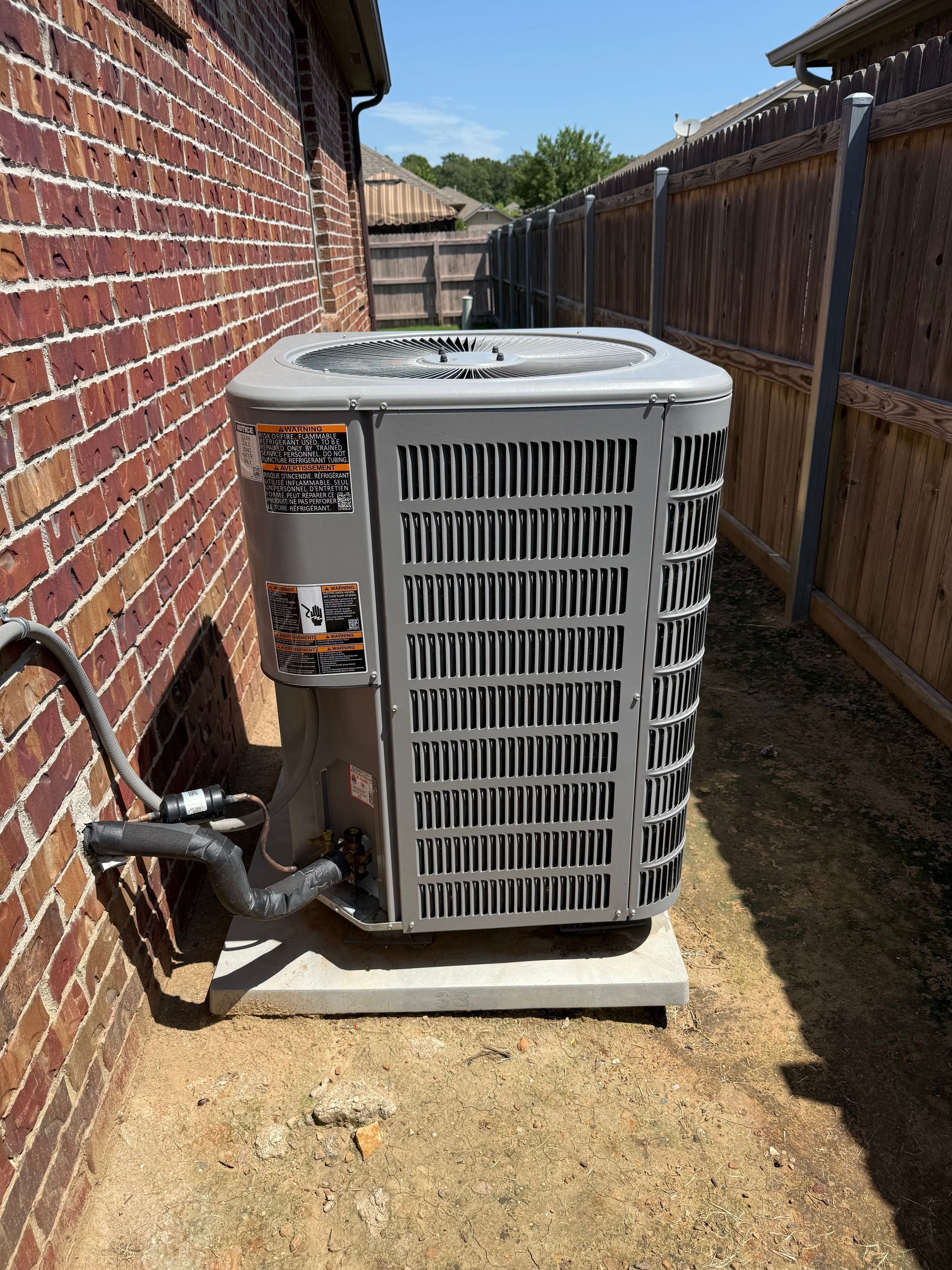 Air conditioning unit on a concrete slab, set between a brick wall and wooden fence, sunny day.