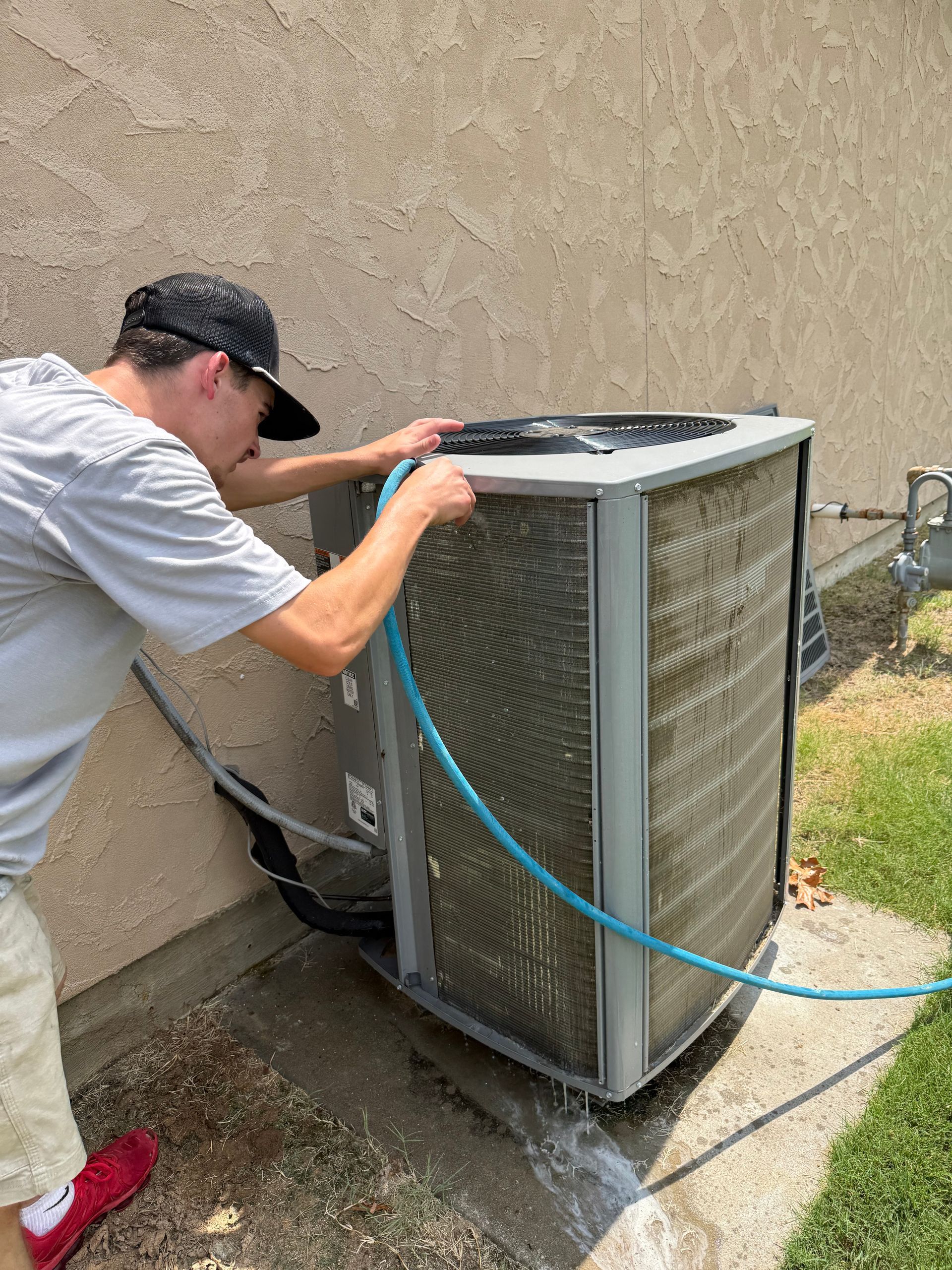 Man hosing down an air conditioning unit outside a building, grass on the right.