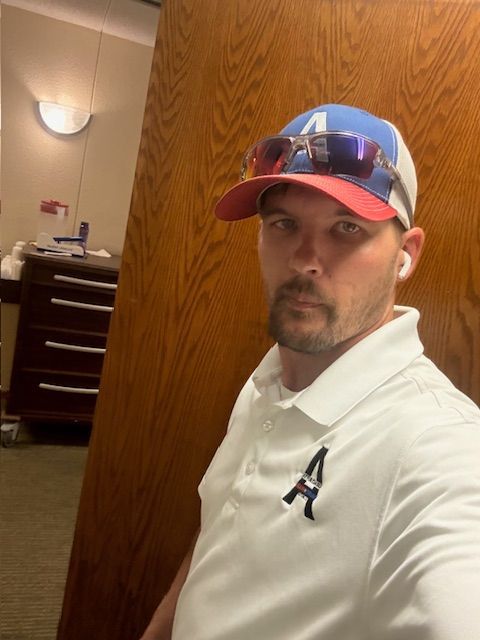 Man wearing a baseball cap, sunglasses, and polo shirt, indoors near a cabinet and wooden wall.
