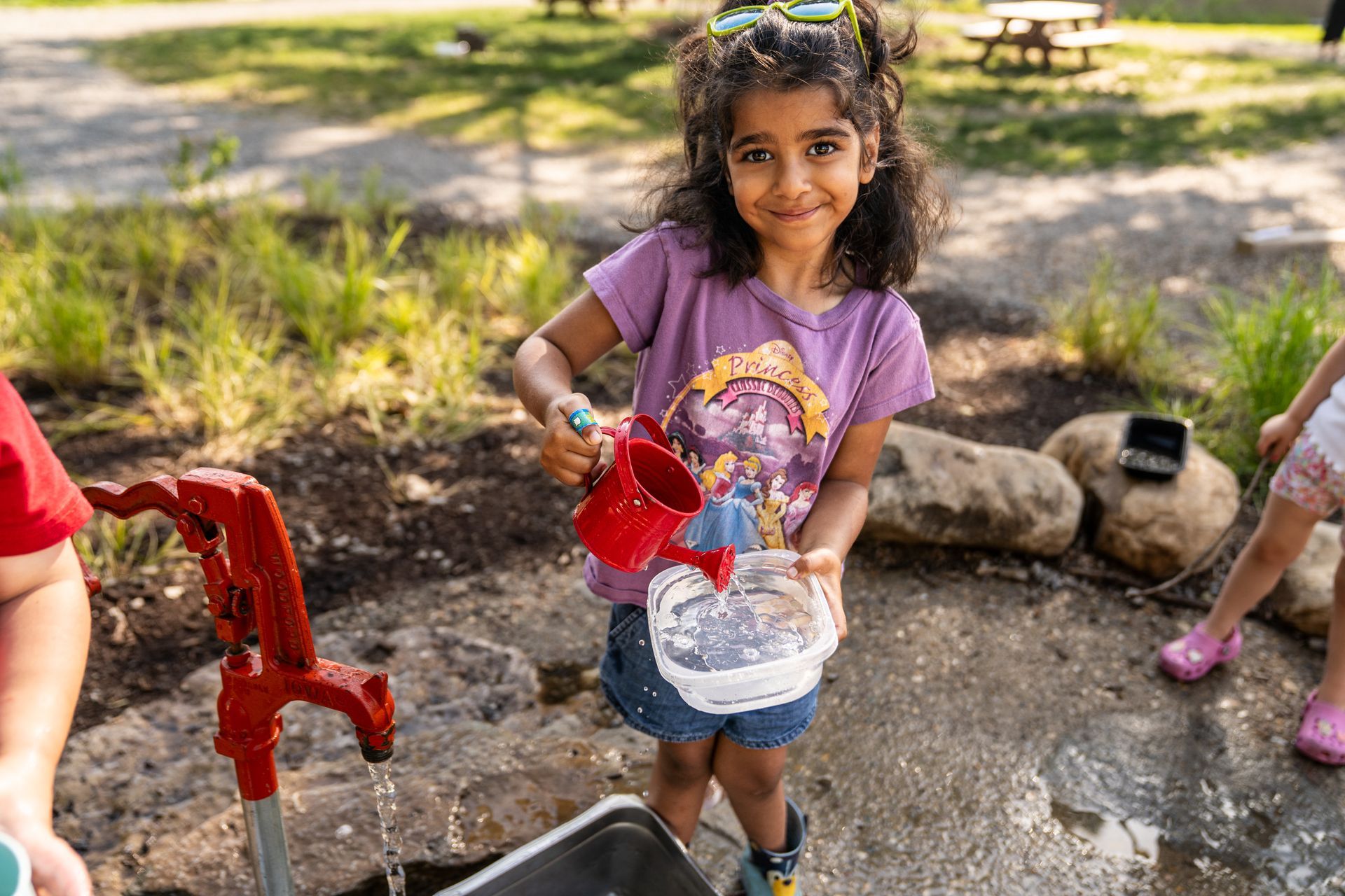 Girl pouring water from a red pitcher into a bowl by a water pump outdoors, smiling.