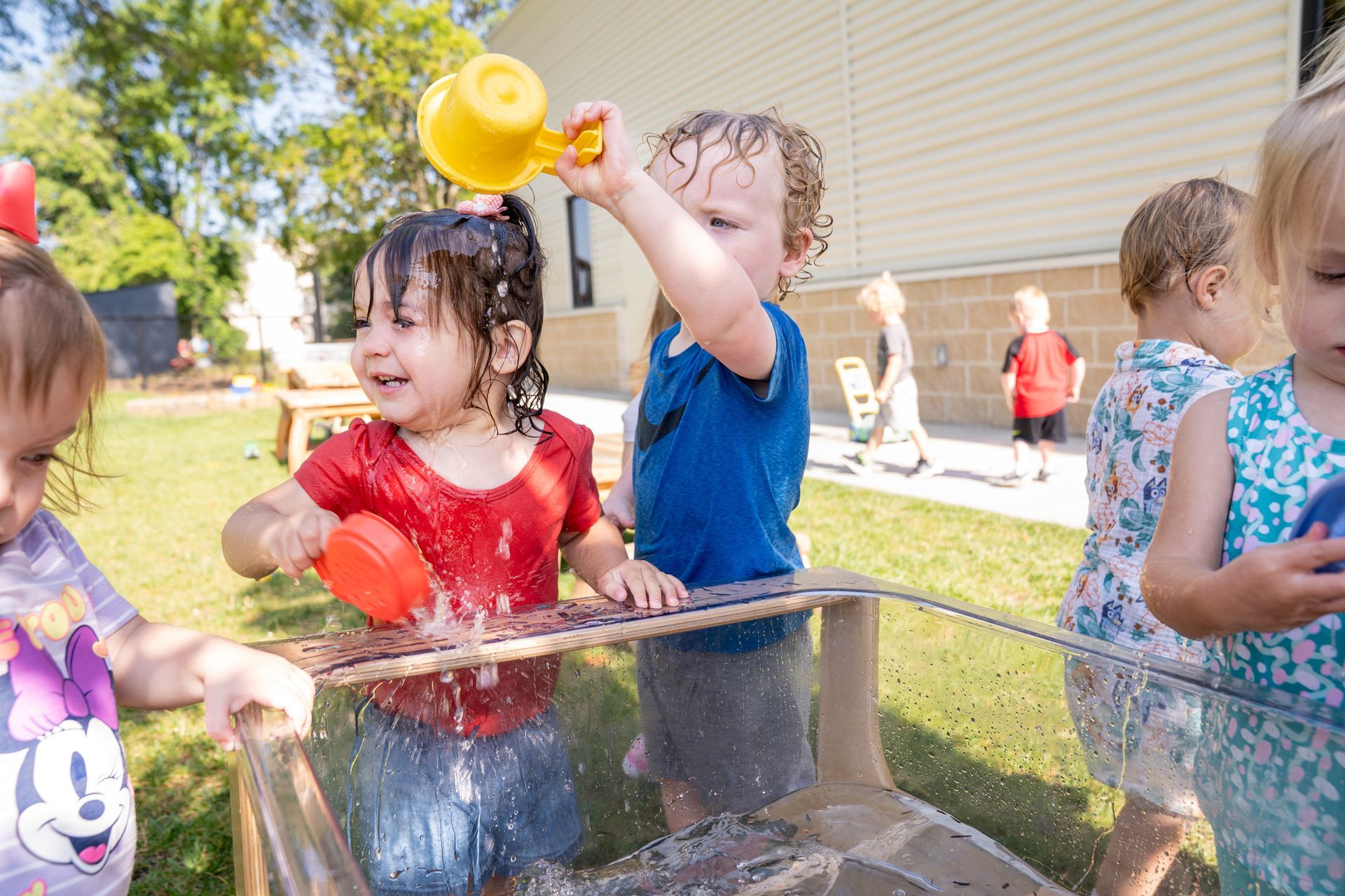 Children playing with water at an outdoor water table. One pours water, others watch and smile.