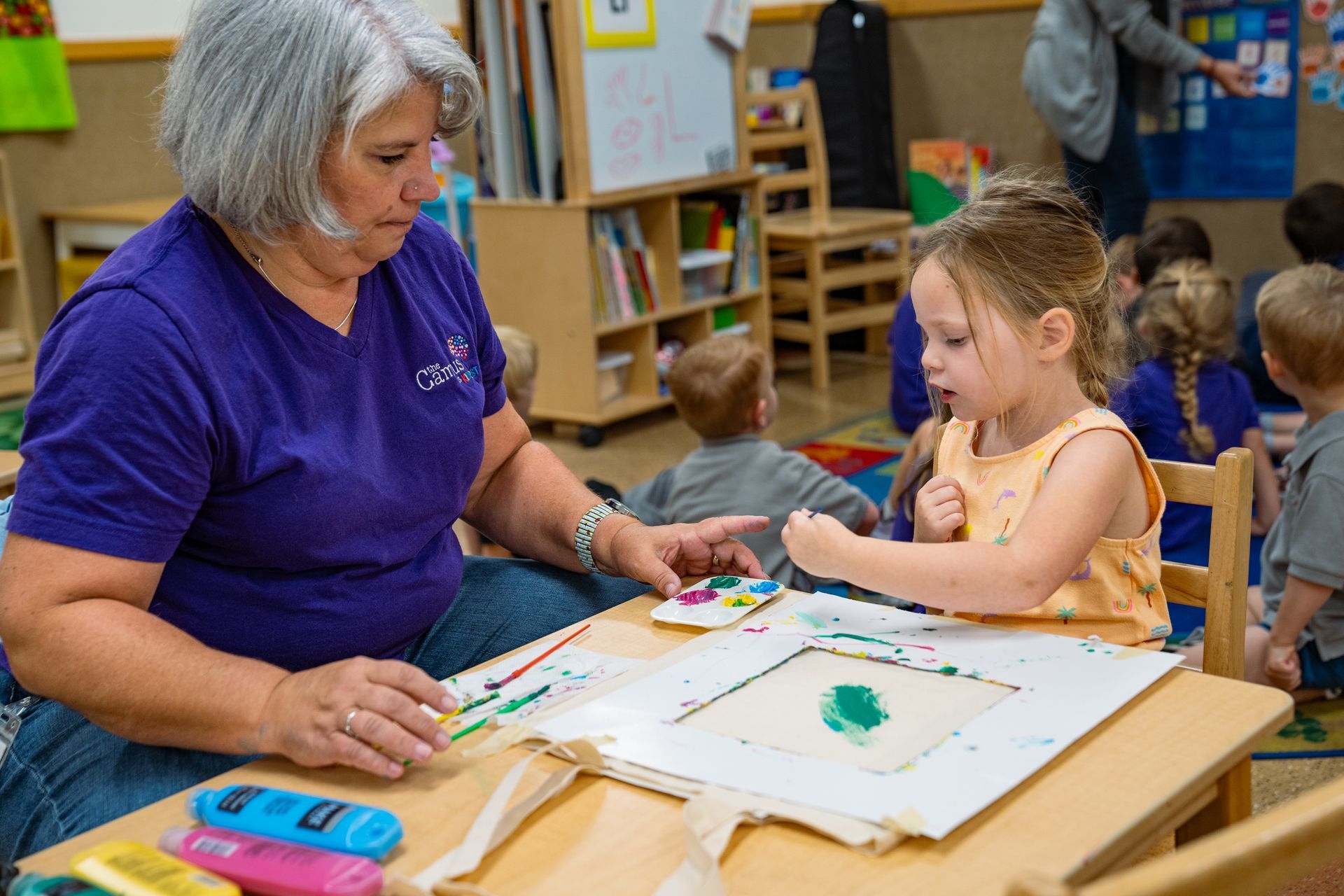 A teacher helps a young girl paint at a table in a colorful classroom.