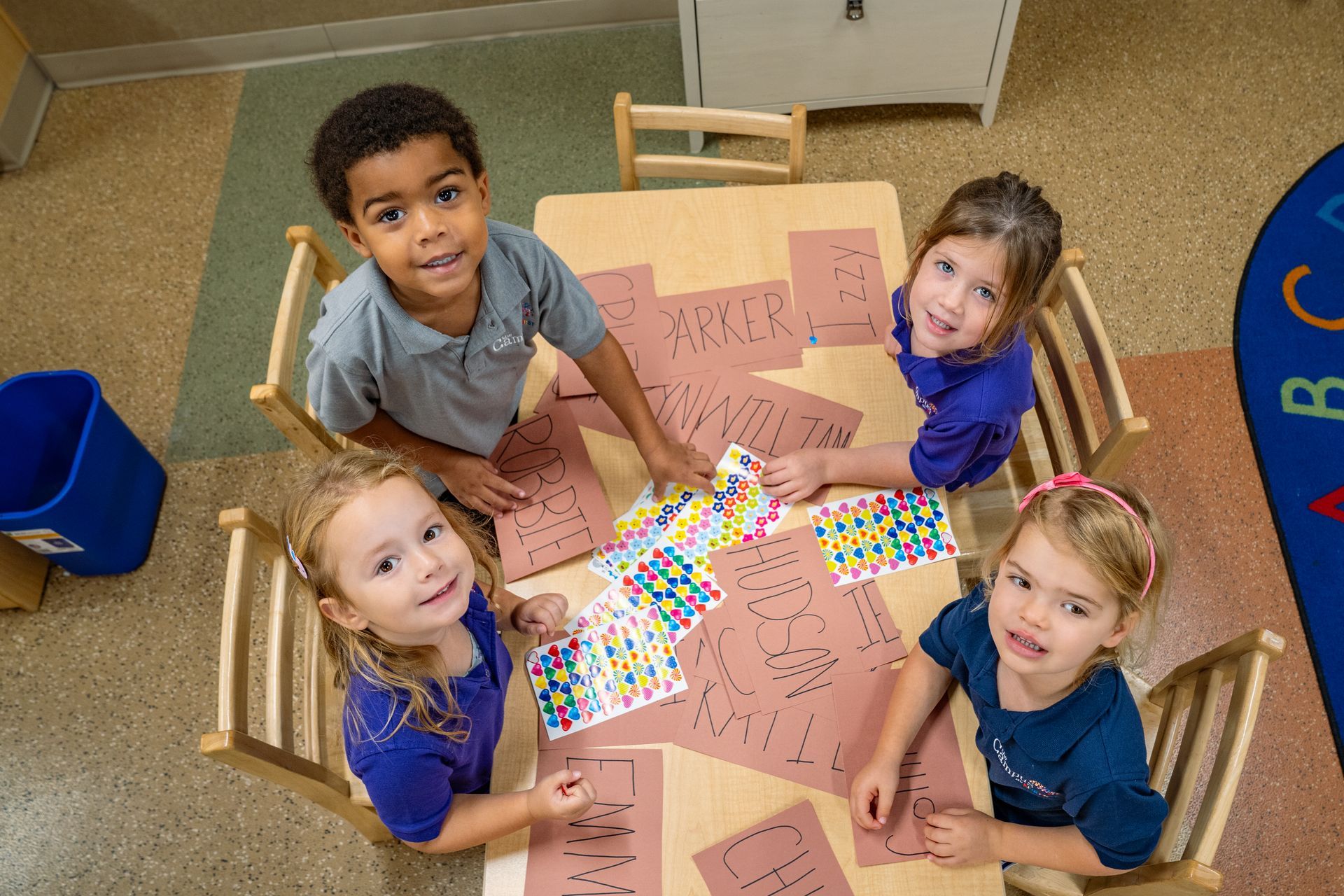Four smiling children around a table, looking up. Papers and stickers on the table in a classroom setting.