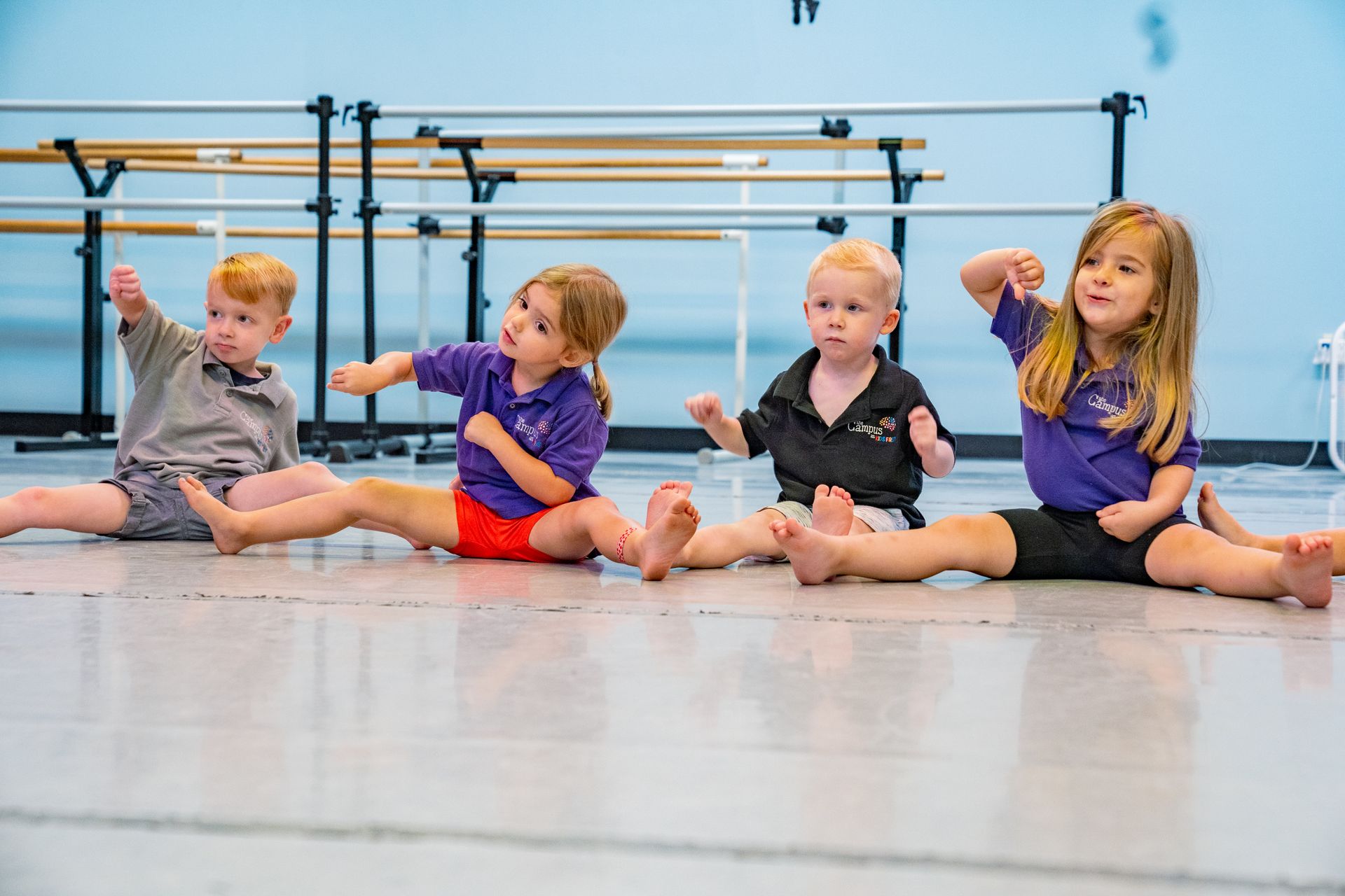 Four young children in a dance studio stretching, arms raised.