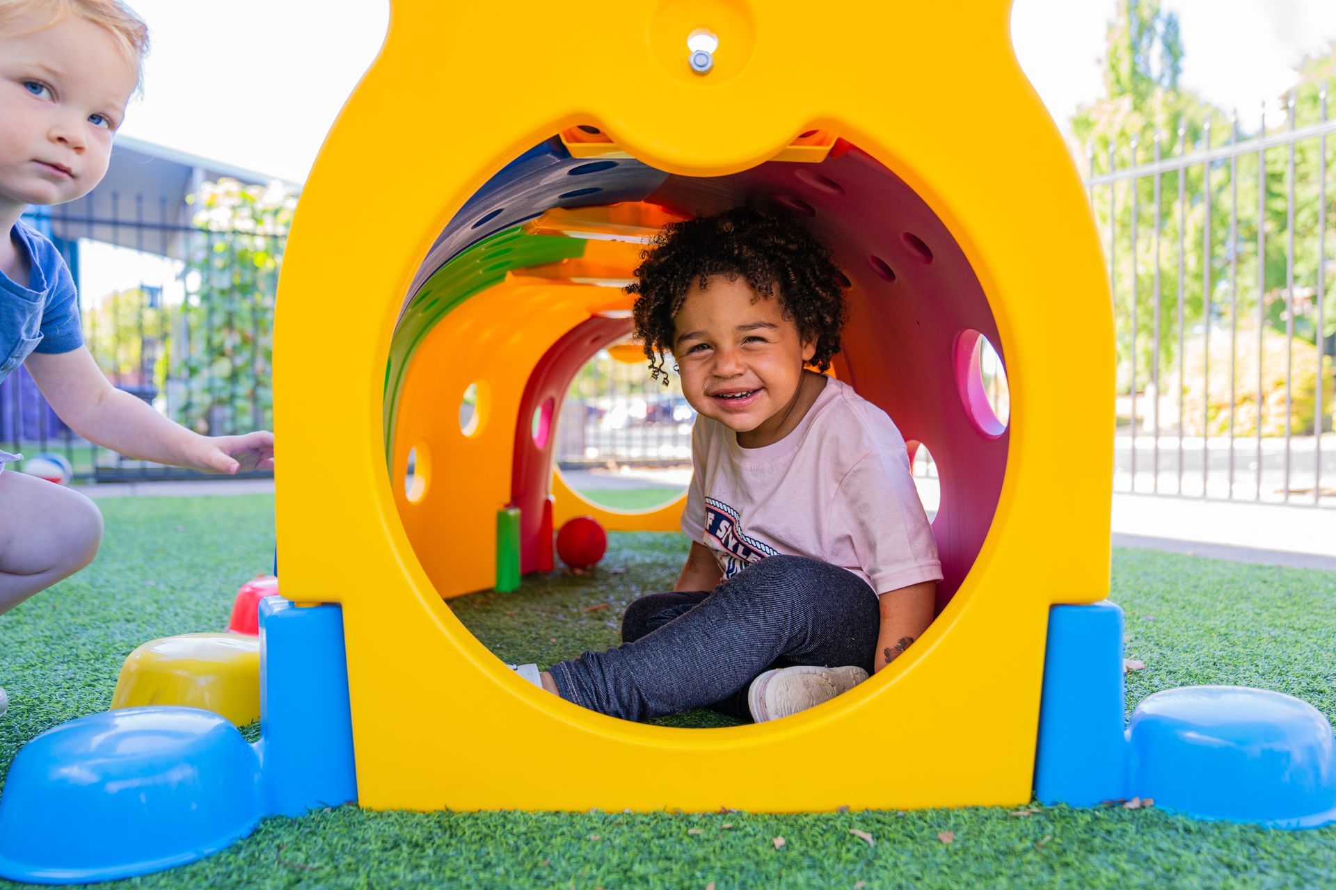 Child smiles from inside a yellow, owl-shaped tunnel on green turf, another child beside.