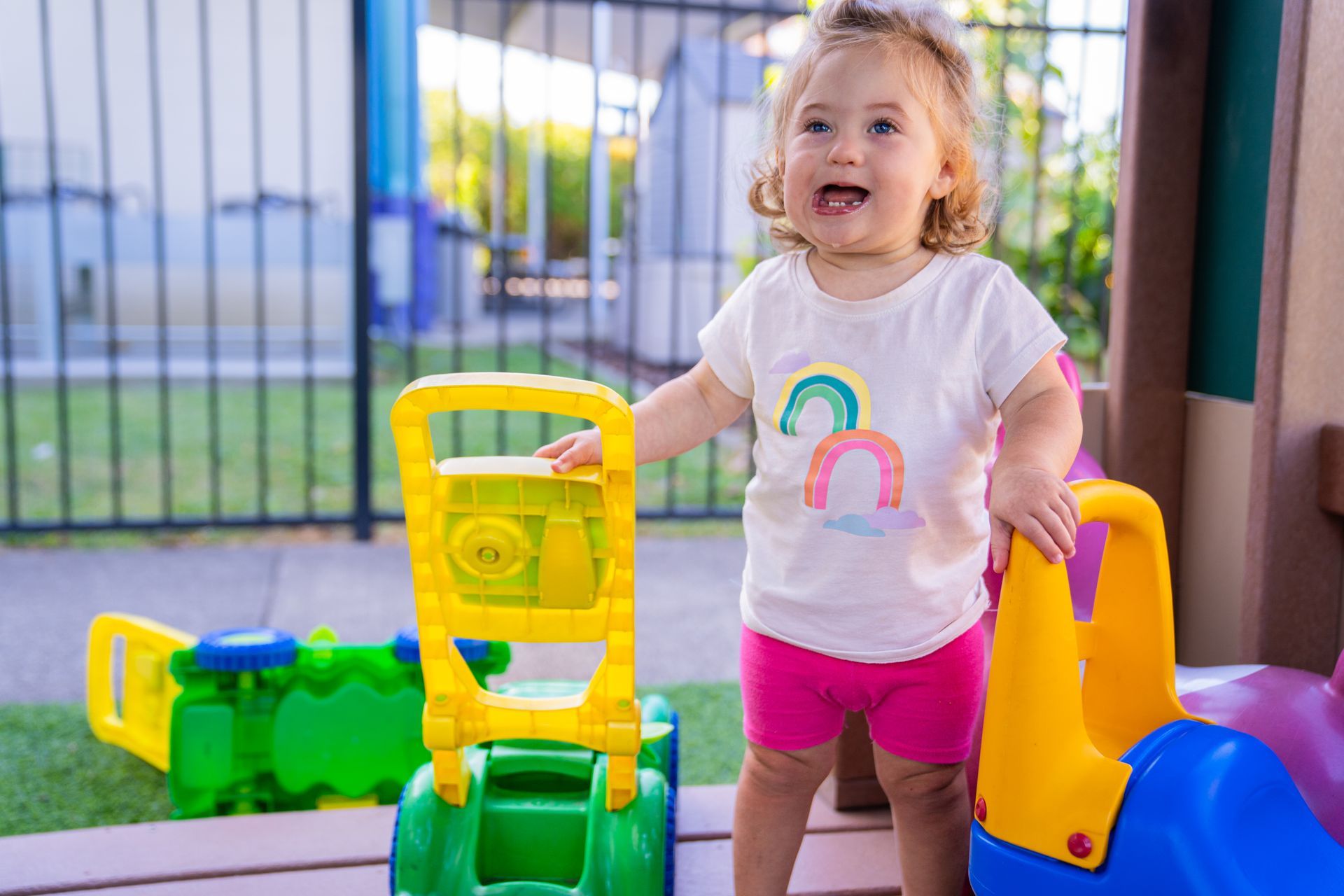 Smiling toddler with Down syndrome pushing a toy lawnmower on a playground.