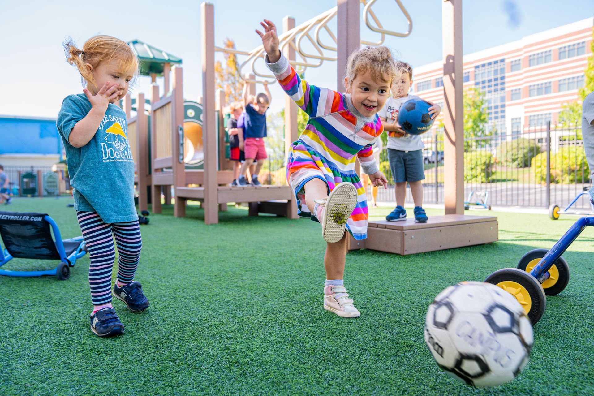 Children playing soccer on a green playground; one kicks the ball, others watch nearby.