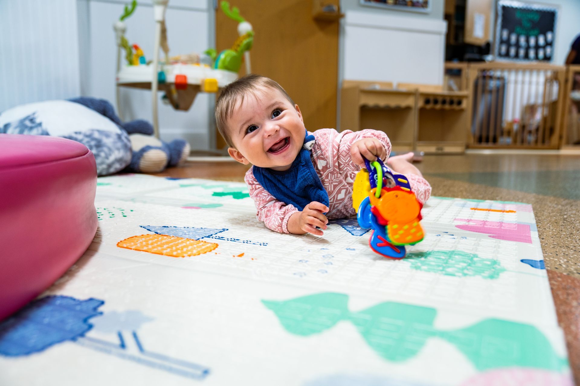 Smiling baby plays on a colorful mat, reaching for a toy. Indoor setting with other toys visible.
