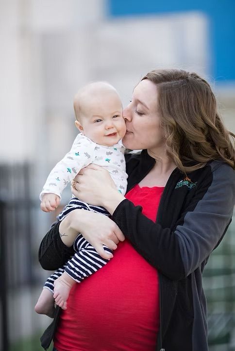Pregnant woman kissing baby, wearing red dress, black jacket; outdoors.