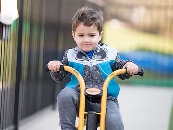 Young boy with brown hair rides a yellow tricycle outdoors, smiling. He wears a blue jacket and gray pants.