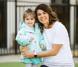 Woman and child smiling and embracing; outdoors. Woman wearing white top, child wearing a blue star jacket.