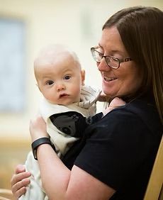 Woman with glasses smiles, holding a baby wearing a hooded onesie.