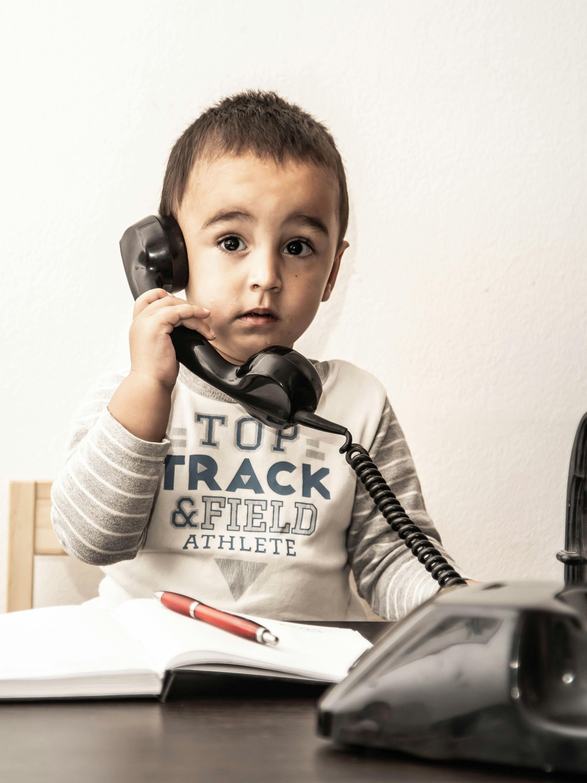 Young child with a serious expression on the phone, sitting at a desk with a notebook and pen.