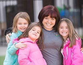 Woman with red hair smiles with three young girls, all smiling, outdoors.