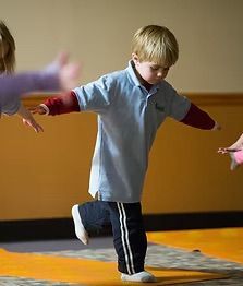 Boy in blue shirt balances on one leg with arms out, indoors.
