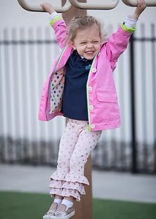 Young girl in pink jacket hangs from monkey bars, smiling.