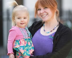 Woman with red hair holding a blonde toddler, both smiling, in front of a blurred background.