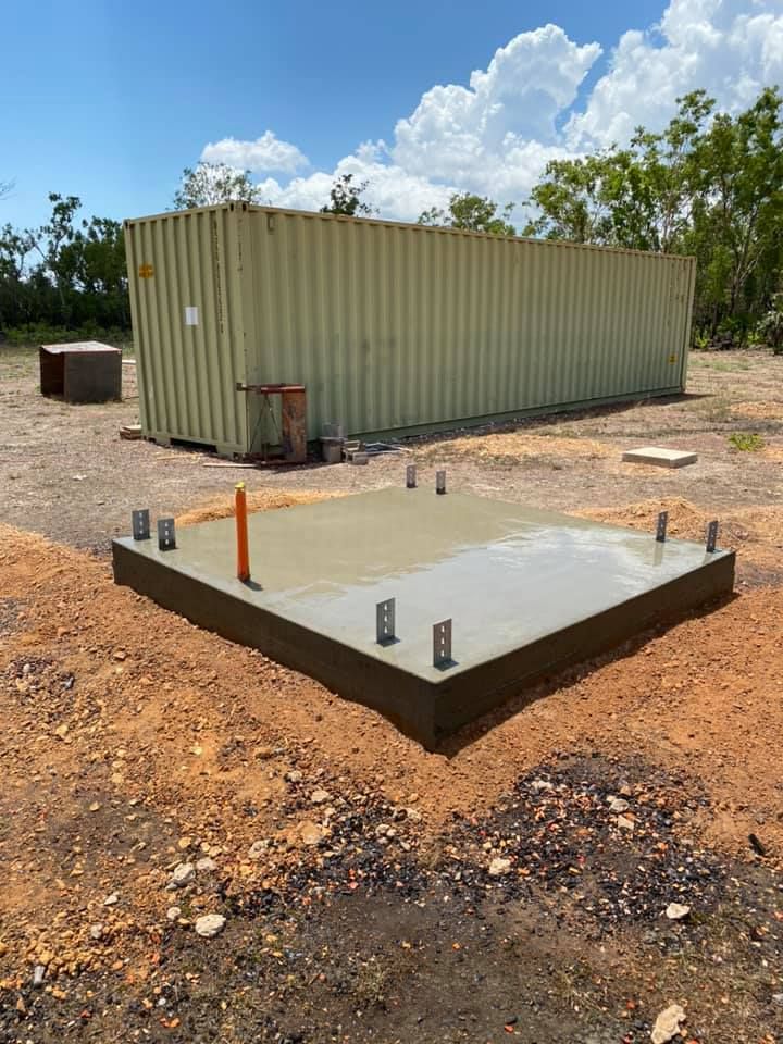 Newly poured concrete pad with bolts, in front of a tan shipping container on a dirt lot, under a blue sky — Dwayne Hamlyn Concreting in Woodroffe, NT