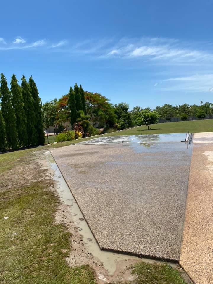Wet concrete patio with water runoff, green grass, and trees under a blue sky — Dwayne Hamlyn Concreting in Woodroffe, NT