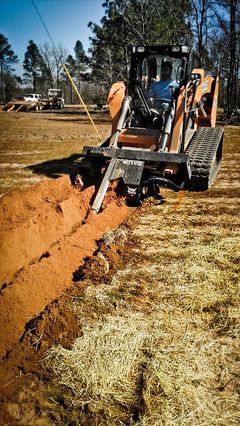Orange trackhoe digging a trench in a field. A person operates the machine. Sunlight illuminates the scene.