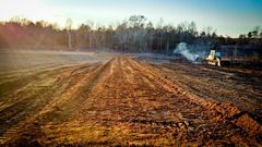 Bulldozer clearing land, brown soil, trees in background, sunny day.