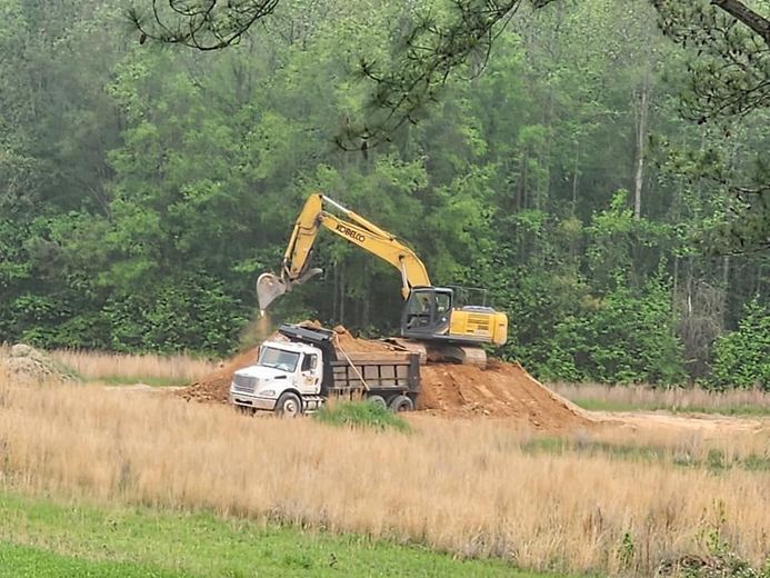 Yellow excavator loading dirt onto a white dump truck, field and trees in the background.