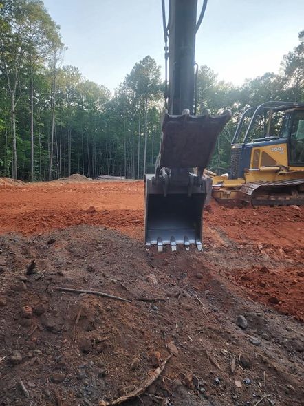 Excavator bucket digging into dirt. Red soil in the background with trees and another machine.
