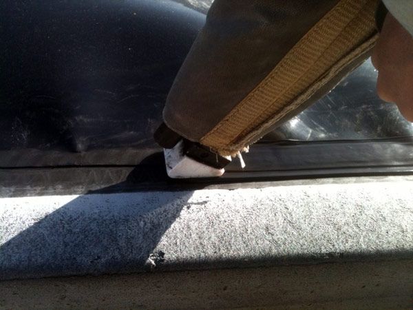 A person applying sealant from a caulk gun to a car window's edge. The caulk gun is beige and brown; the car is black, with concrete nearby.