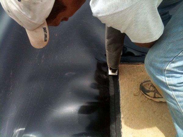 Person applying sealant to a dark, curved surface, likely a container, wearing a white hat and jeans.