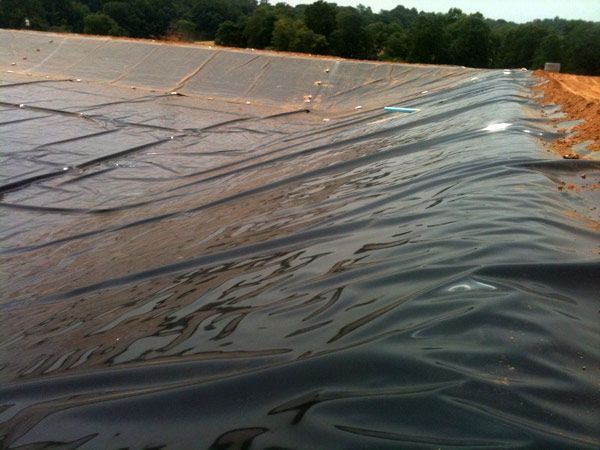 Large black plastic liner covering the interior of a dirt-walled containment structure, likely for water or waste, under a sunny sky.