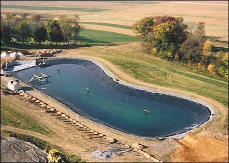 An aerial view of a large lined lagoon used for wastewater treatment, surrounded by fields and trees.