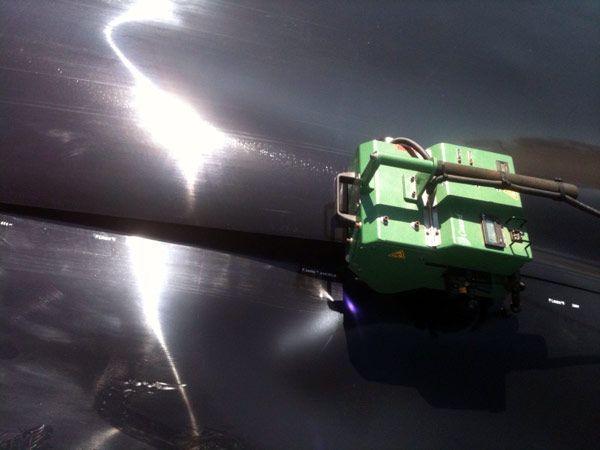 A green robotic device welding on a shiny, dark surface, likely outdoors. Bright sunlight reflects off the surface.