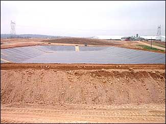 A large, rectangular lagoon covered in black lining, with a dirt berm in the foreground and agricultural buildings in the distance.