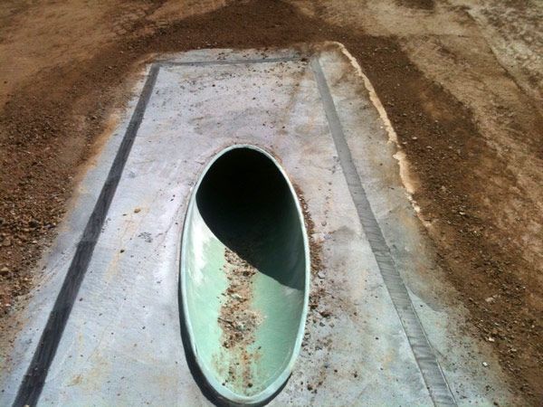 Oval-shaped drainage culvert, pale green inside, set into concrete with dirt surroundings.